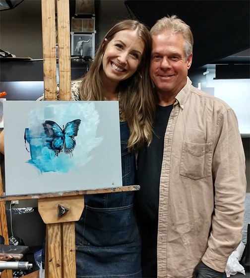Two people posing with a painting of a blue butterfly on an easel.