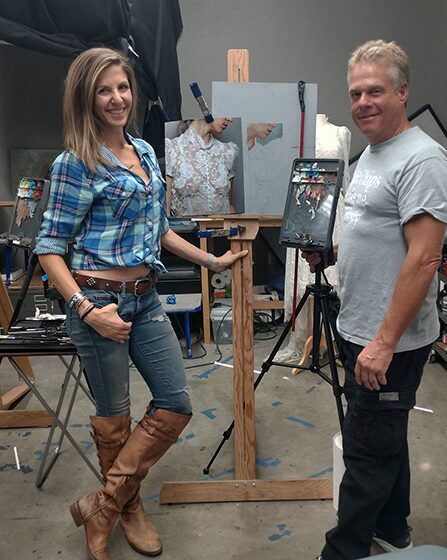 Two artists posing beside an easel with a painting in progress.
