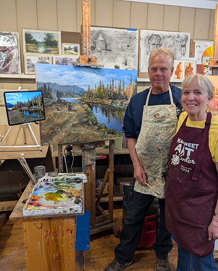 Two artists posing with landscape paintings in an art studio.