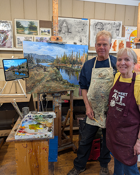 Two artists posing with landscape paintings in an art studio.